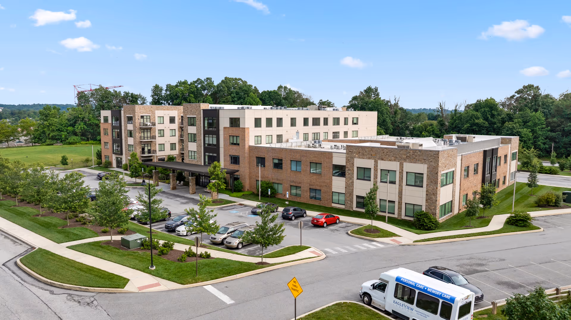 Exterior view of a multi-story senior living building with a parking lot, landscaped grounds, and a shuttle van.
