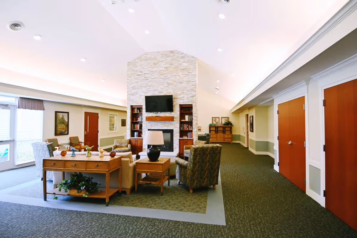 A spacious and well-lit common area with a high ceiling, featuring a stone fireplace with a mounted TV above it. Comfortable armchairs and sofas are arranged around the fireplace, with side tables and a console table decorated with small sculptures and a lamp. The room has carpeted floors, large windows letting in natural light, and wooden doors along the hallway.