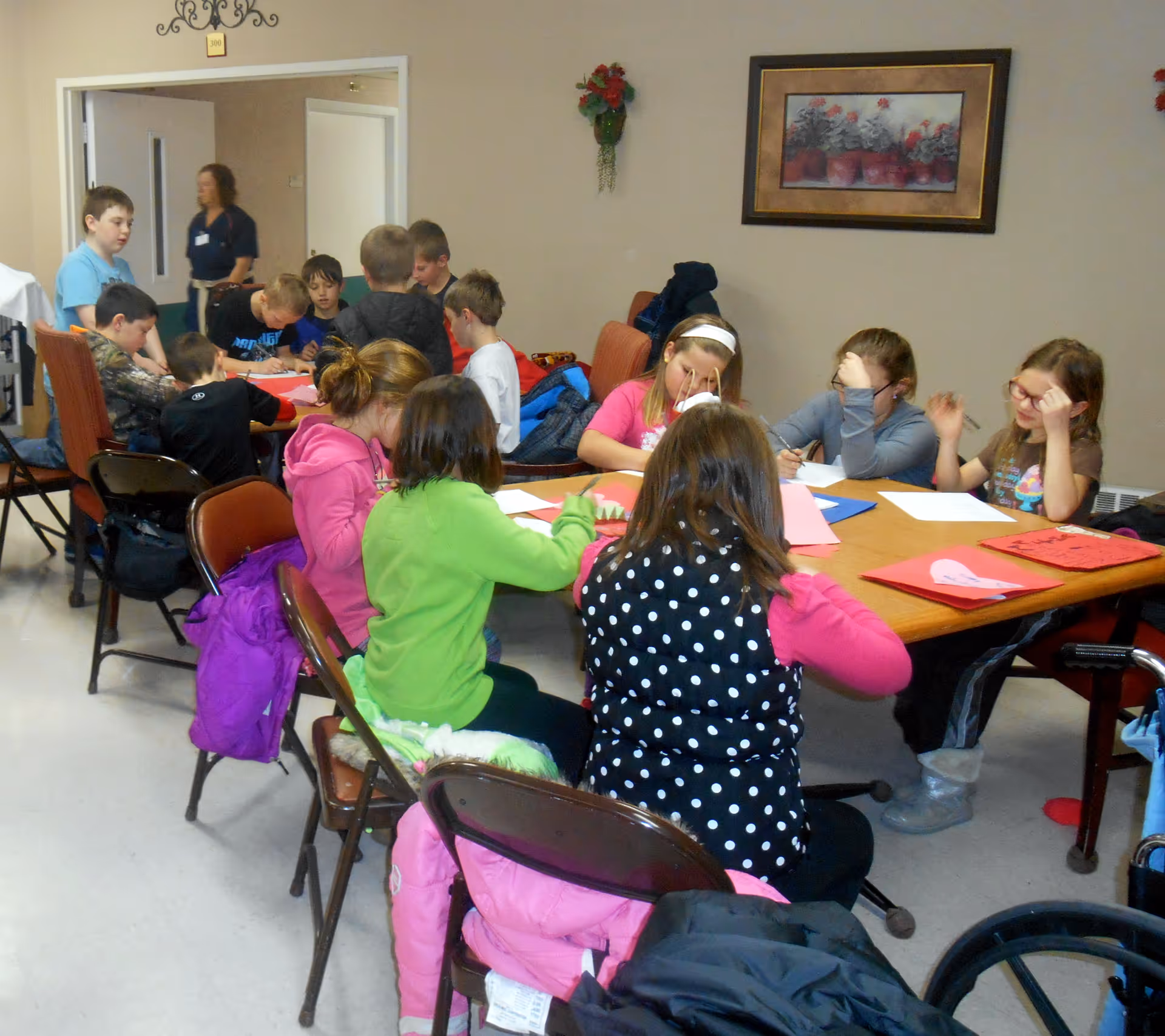 A group of children sitting around a table engaged in drawing or writing activities inside a room. There are chairs around the table, and a woman is standing in the background near an open door. The walls are decorated with a framed picture and a small floral arrangement.