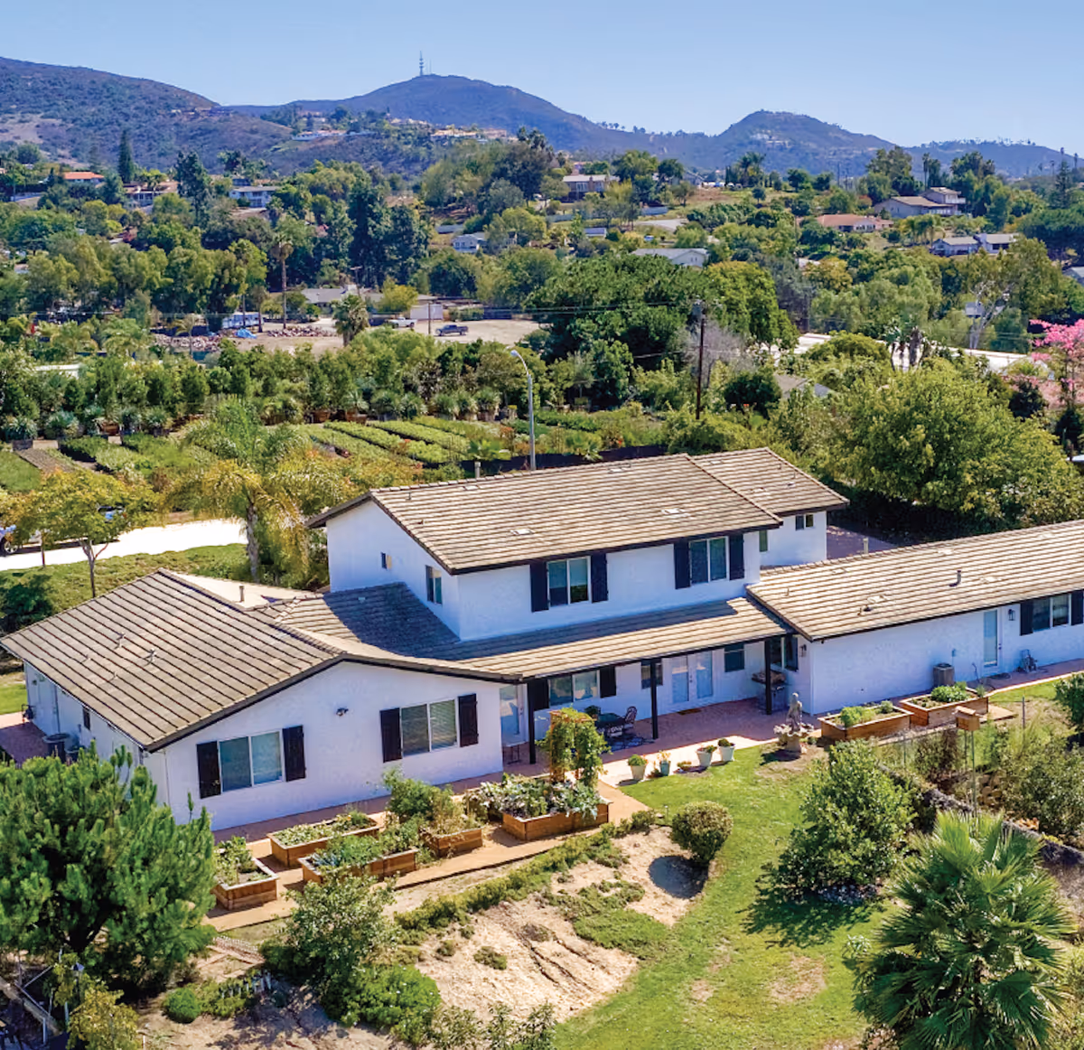 Aerial view of a white two-story building with a tiled roof surrounded by greenery, garden beds, and trees, set against a backdrop of hills and a clear blue sky.