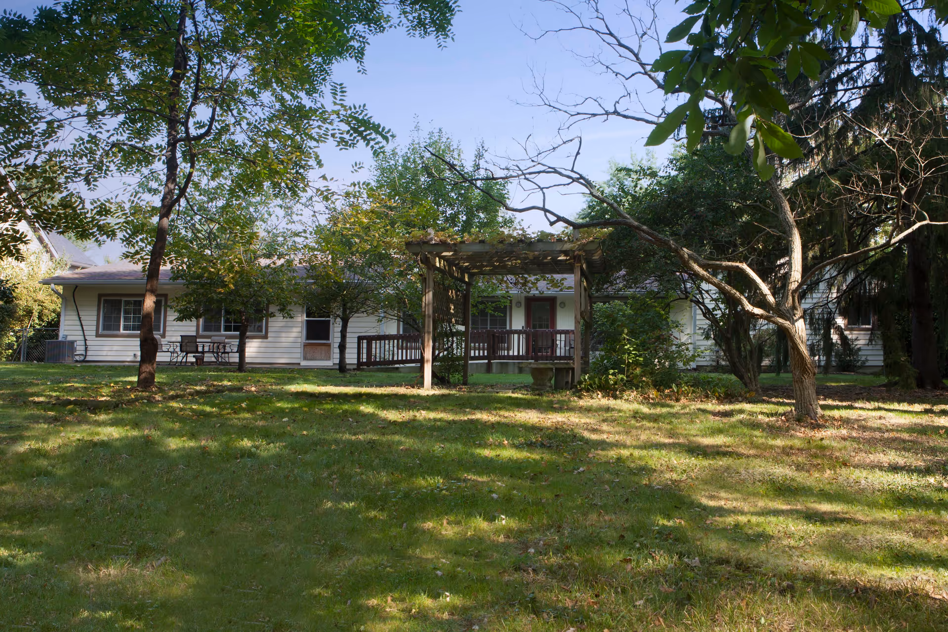 A grassy outdoor area with scattered trees and a wooden pergola in the center. In the background, there is a single-story building with a porch and outdoor seating. The scene is shaded with sunlight filtering through the trees.