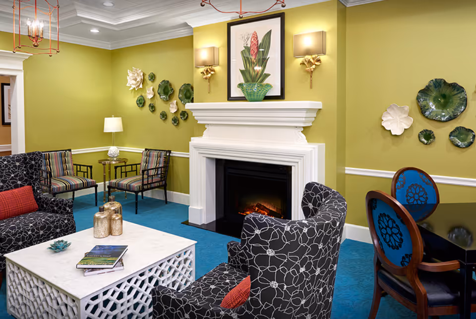 A cozy living room area with green walls and blue carpet. The room features a white fireplace with a framed botanical print above it, flanked by two wall sconces. There are patterned armchairs with red accent pillows, a white lattice coffee table with books and decorative items, and a small seating area with striped chairs and a side table. Decorative ceramic wall art is displayed on the walls.