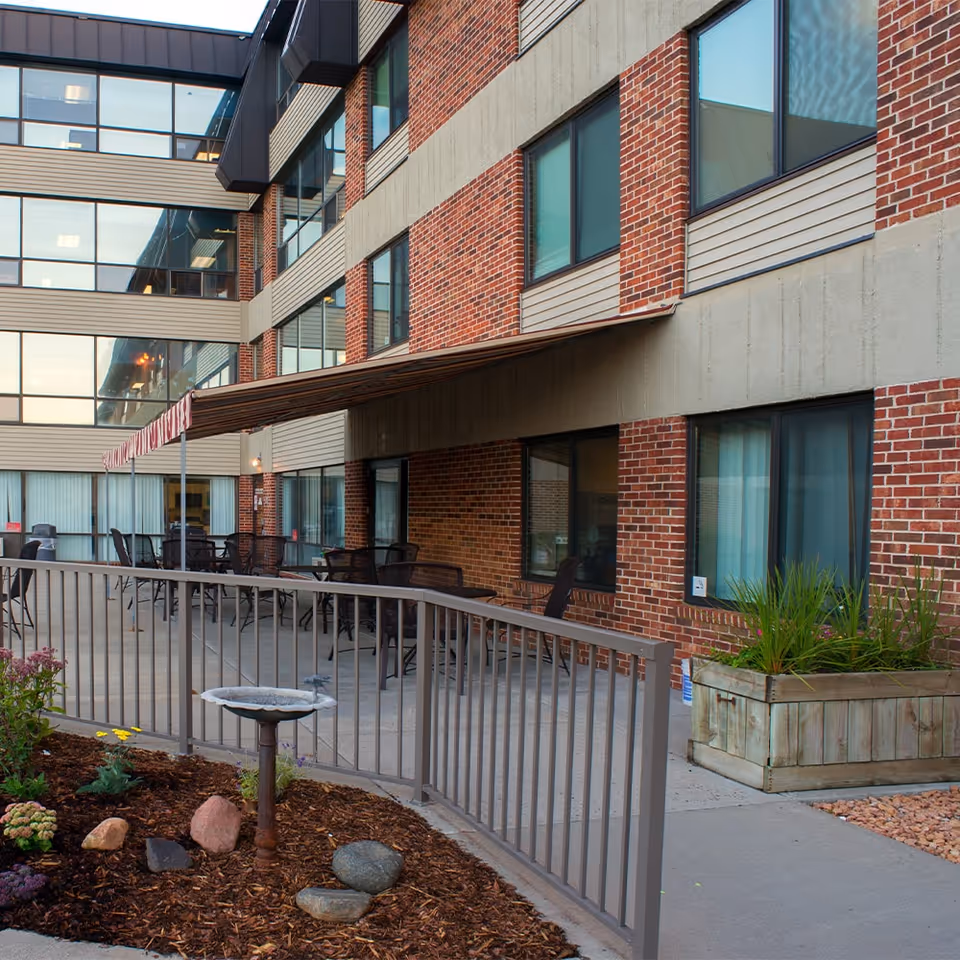 Outdoor patio area with tables, chairs, planters and a metal railing in front of a multi-story brick building.
