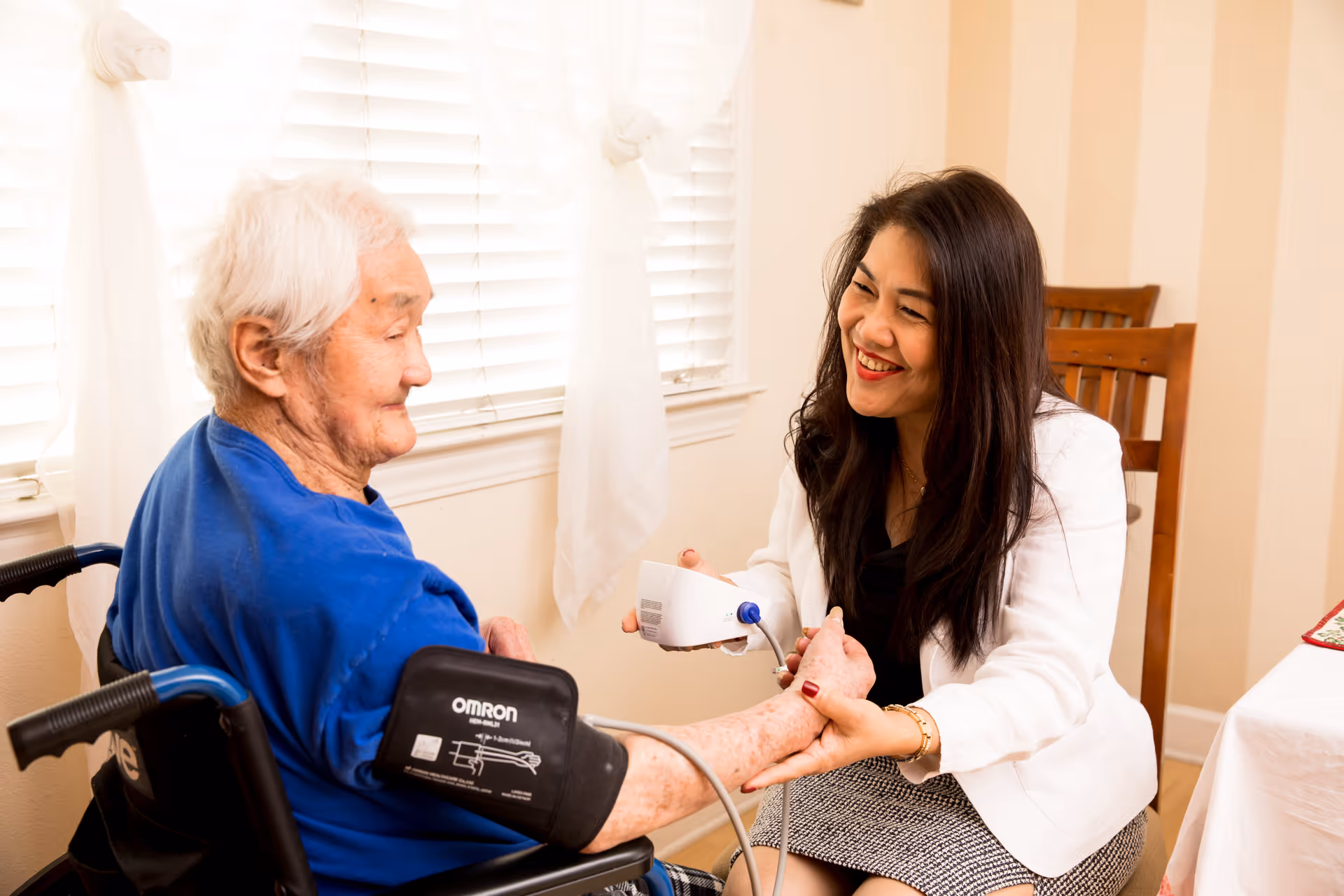 A healthcare professional in a white coat is smiling and taking the blood pressure of an elderly person seated in a wheelchair inside a well-lit room with white curtains and wooden chairs.