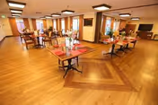 A spacious dining room with wooden floors and multiple tables set with red placemats and condiments. The room is well-lit with ceiling lights and has large windows with curtains allowing natural light to enter. There is a television mounted on the wall and a fireplace in the background.