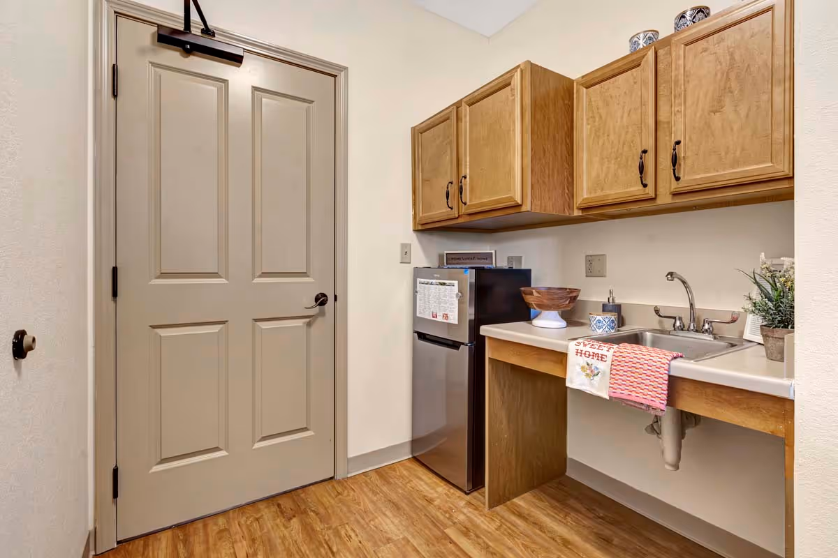 Small kitchenette with wooden cabinets, a sink, a mini refrigerator, and a closed beige door.
