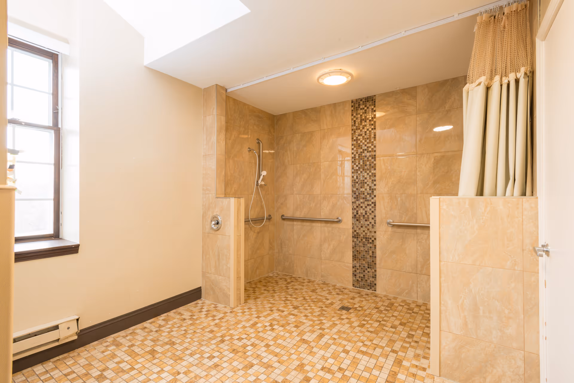 A spacious, tiled shower area with beige and brown mosaic tiles on the floor and walls. The shower has two grab bars, a handheld showerhead, a skylight above, and a window to the left. There is a curtain on the right side for privacy.