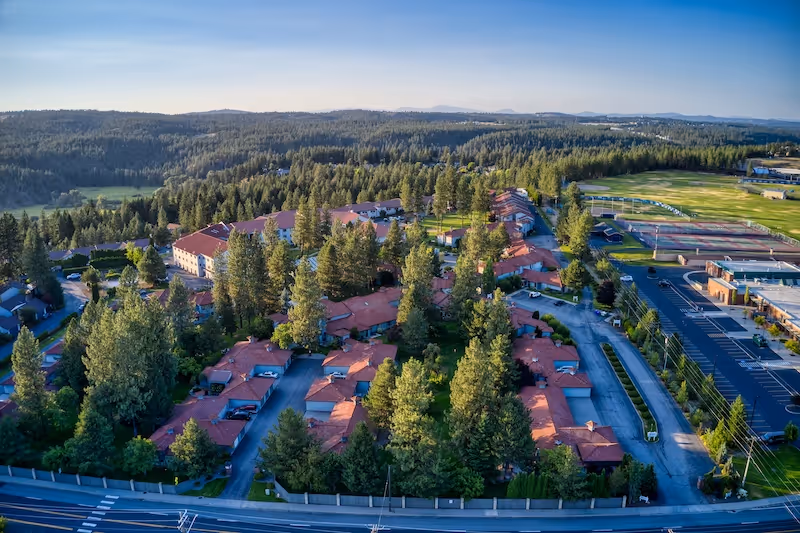 Aerial view of Fairwood Retirement Village & Assisted Living showing multiple buildings with red roofs surrounded by tall trees and greenery. There is a parking lot and sports fields visible on the right side, and a forested landscape extends into the distance under a clear sky.