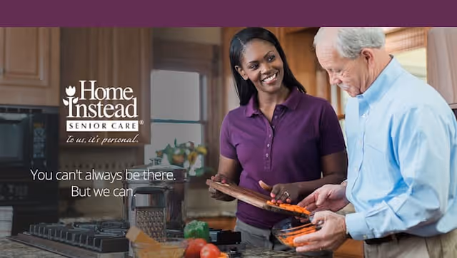 A smiling woman in a purple shirt helps an elderly man in a blue shirt prepare sliced carrots in a kitchen setting. The background shows kitchen cabinets and a window with natural light.