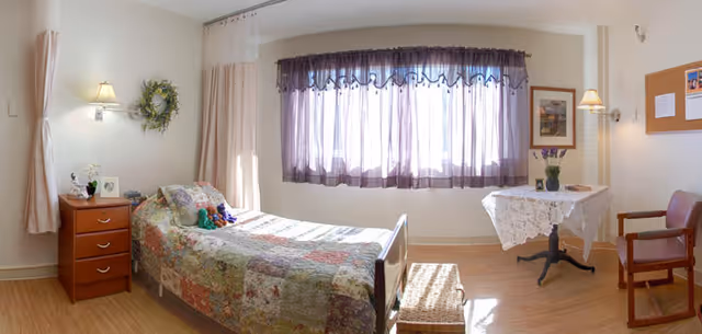 A bright and cozy nursing facility bedroom with a single bed covered in a colorful patchwork quilt. Next to the bed is a wooden nightstand with a lamp, a small plant, and a framed photo. A wreath hangs on the wall above the nightstand. Across from the bed is a small table with a lace tablecloth, a vase of flowers, and a chair beside it. The room has large windows with sheer purple curtains allowing natural light to fill the space.