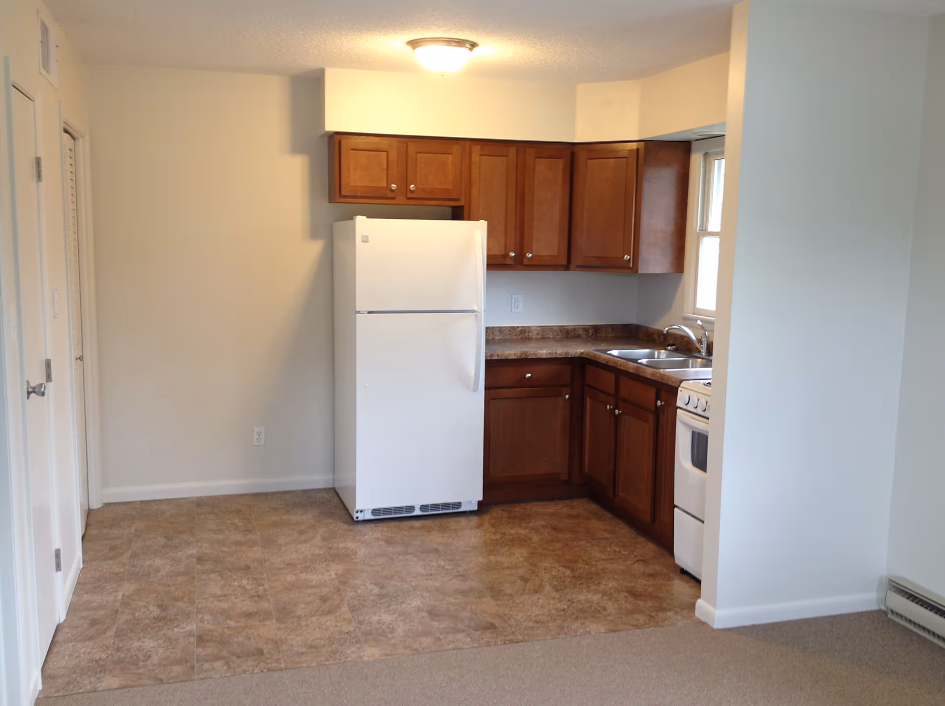 A small kitchen area with brown wooden cabinets, a white refrigerator, a white stove, and a double sink under a window. The floor is tiled in the kitchen area and carpeted in the adjacent space. The walls are painted white and there is a ceiling light fixture.