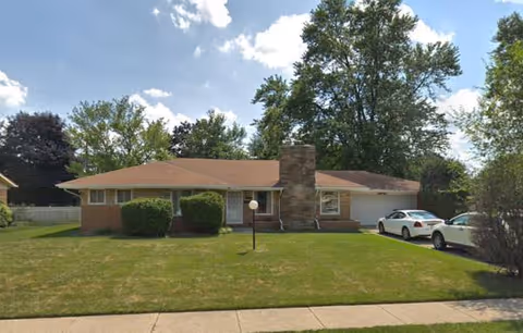 Single-story ranch-style senior living facility with a manicured lawn, driveway, and parked cars.