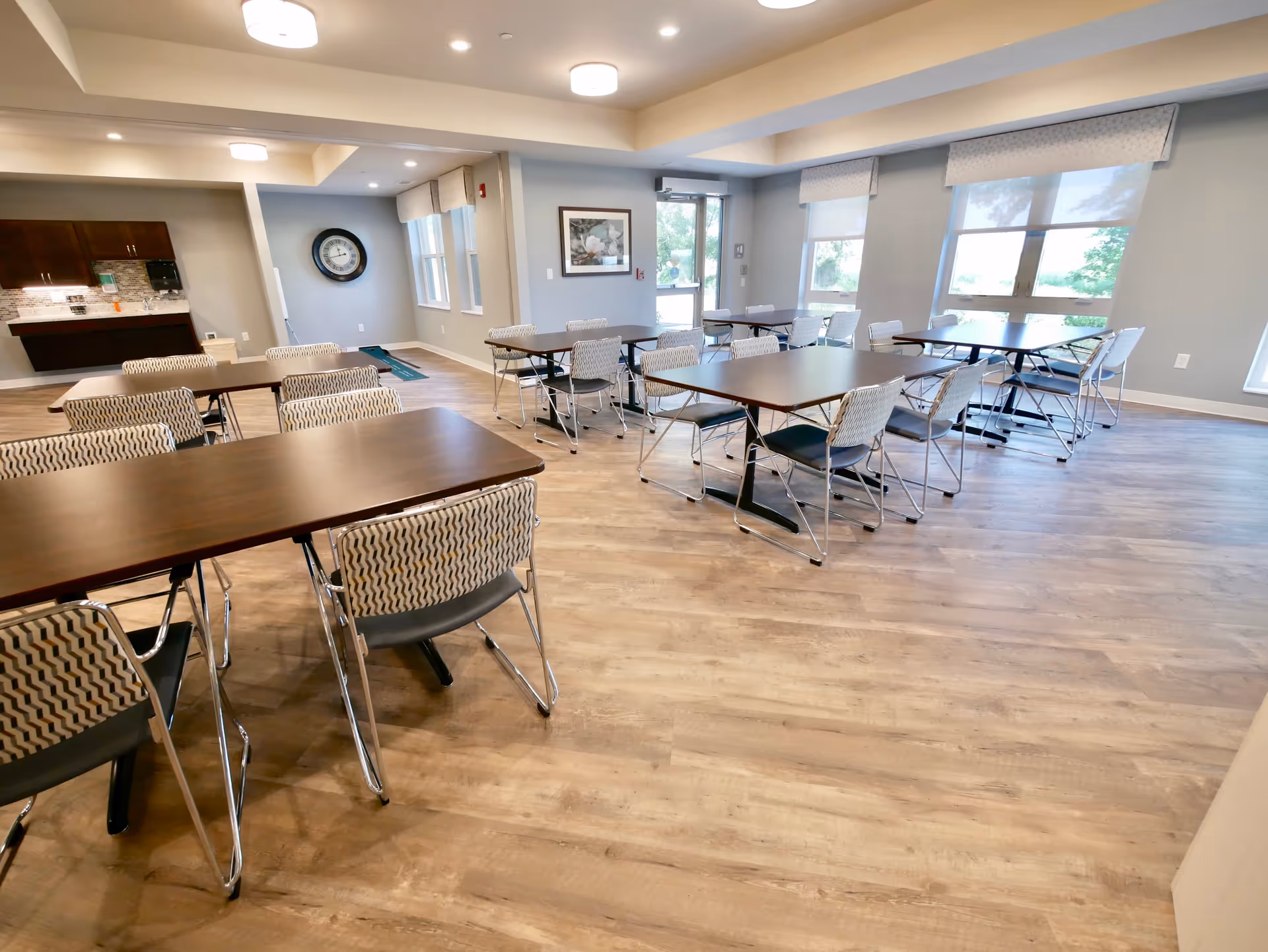 A spacious dining room with multiple rectangular wooden tables and patterned chairs arranged neatly. The room has large windows with light-colored shades, light wood flooring, and soft gray walls. There is a kitchenette area with dark wooden cabinets and a clock on the wall in the background.
