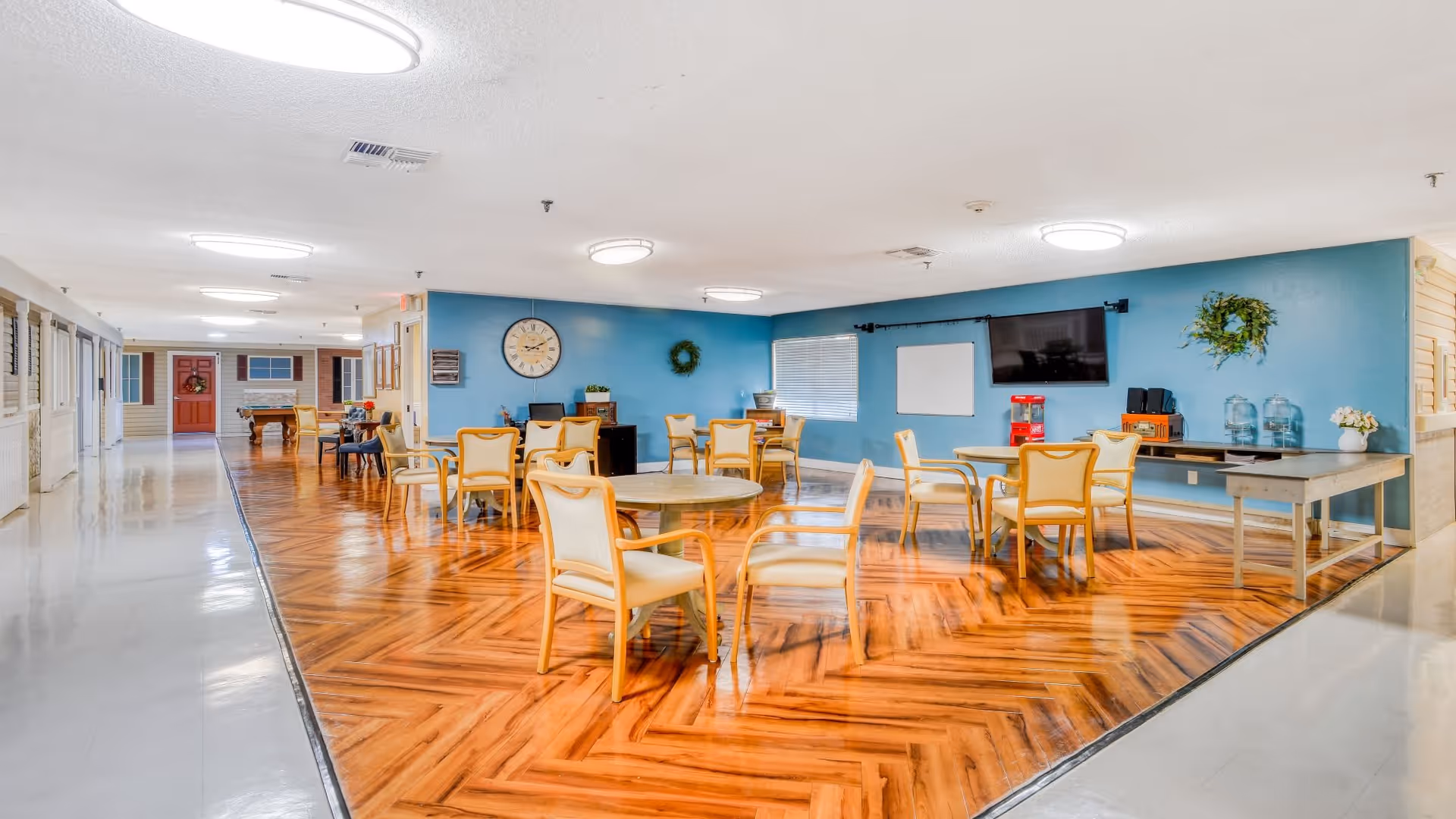 A spacious common area in an assisted living facility with several round tables and chairs arranged on a polished wooden floor. The walls are painted blue and decorated with a large clock, wreaths, a flat-screen TV, and a whiteboard. There is a popcorn machine and other small appliances on a long table against the wall. The area is well-lit with ceiling lights, and a hallway with doors and windows is visible to the left.