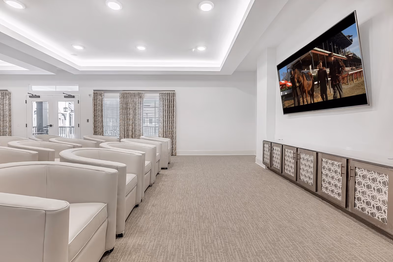 A modern media room with rows of white cushioned armchairs facing a large flat-screen TV mounted on a white wall. Below the TV is a long cabinet with decorative panel doors. The room has recessed ceiling lights, beige carpet, and windows with patterned curtains.