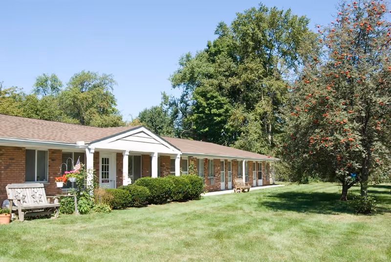 Single-story brick building with a covered entrance and several doors and windows, surrounded by green grass, bushes, and trees under a clear blue sky.