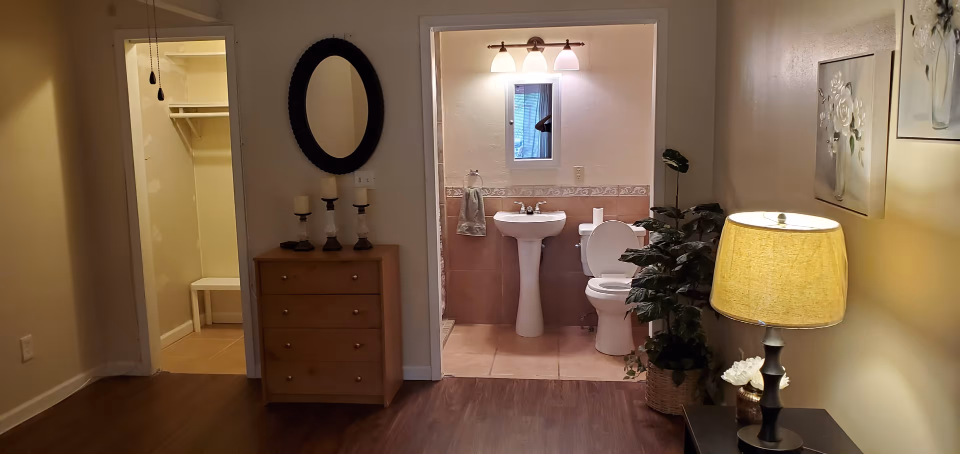 Interior view of a room in Morning Glory Assisted Living showing a small wooden dresser with three candles and a round mirror above it. To the right, there is a bathroom with a pedestal sink, toilet, and a small window above the sink. A potted plant is placed near the bathroom entrance. On the right side of the image, there is a table with a lamp and wall art featuring floral designs. To the left, there is an open doorway leading to a closet with a bench inside.
