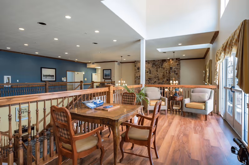 A bright and spacious interior common area with wooden flooring, a wooden table with four chairs, two cushioned armchairs with yellow seat cushions, a small round side table with a plant, stone accent wall, and large windows with yellow curtains allowing natural light to fill the space.