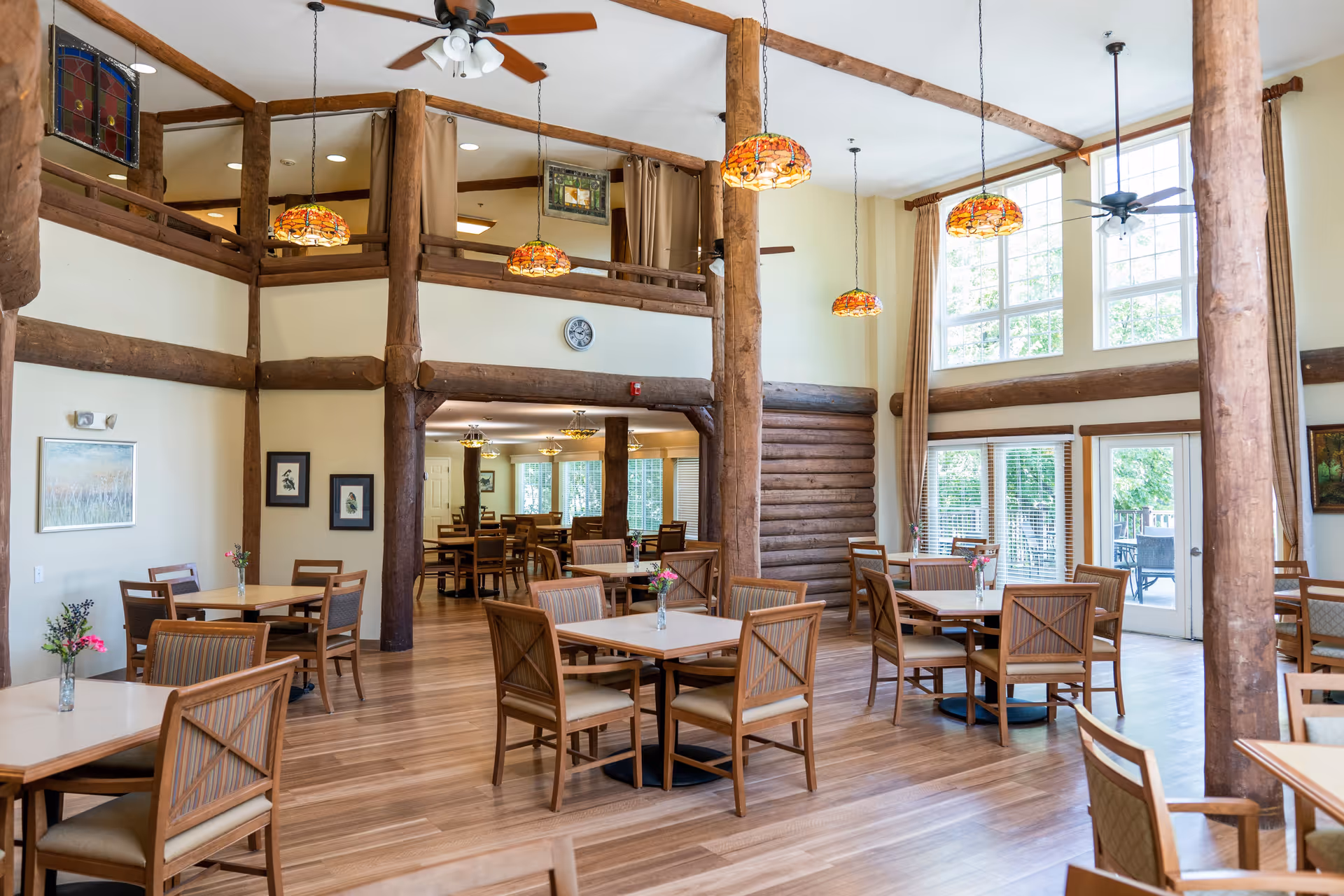 Spacious dining room with wooden tables and chairs, large windows letting in natural light, wooden beams and columns, and hanging decorative pendant lights. The room has a high ceiling with ceiling fans and light wood flooring.