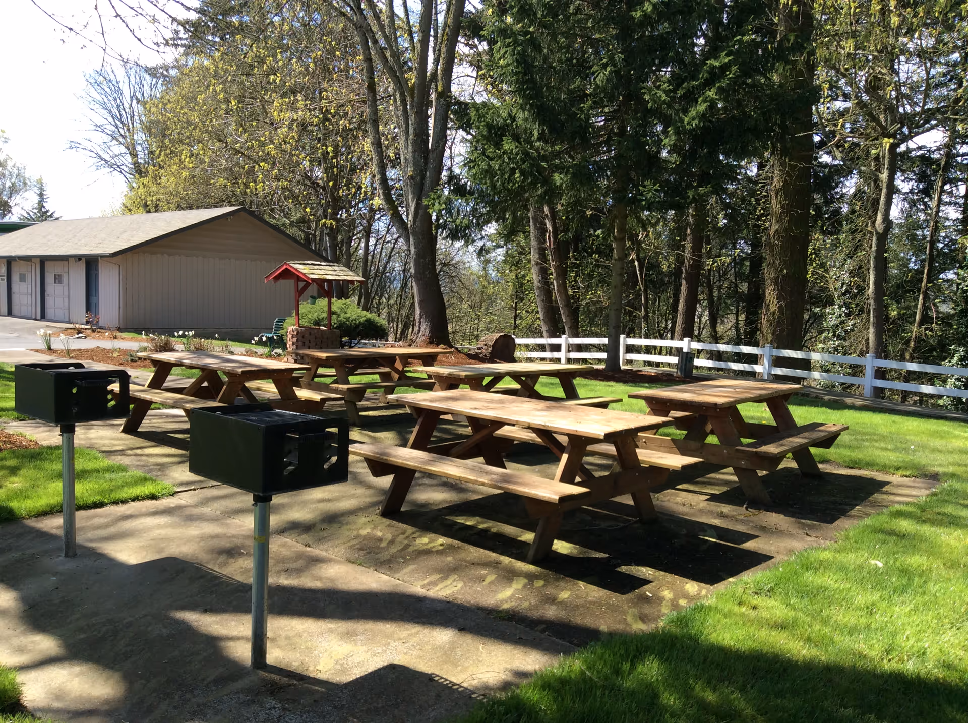 Outdoor picnic area with several wooden picnic tables and two black metal grills on concrete pads, surrounded by green grass, trees, and a white fence. A small building is visible in the background.