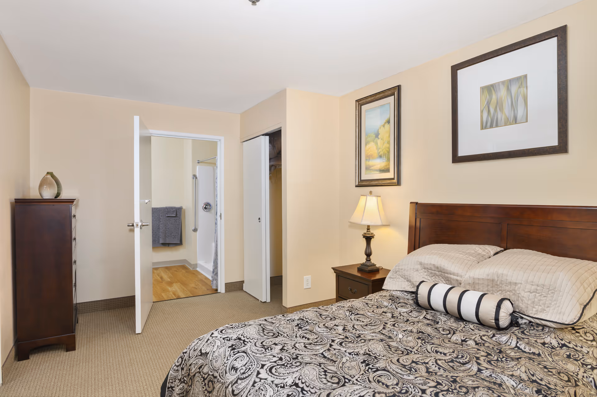 A bedroom in a senior living facility with a bed featuring a patterned bedspread and pillows, a wooden nightstand with a lamp, framed artwork on the walls, a tall dresser, an open door leading to a bathroom, and a closet with sliding doors.
