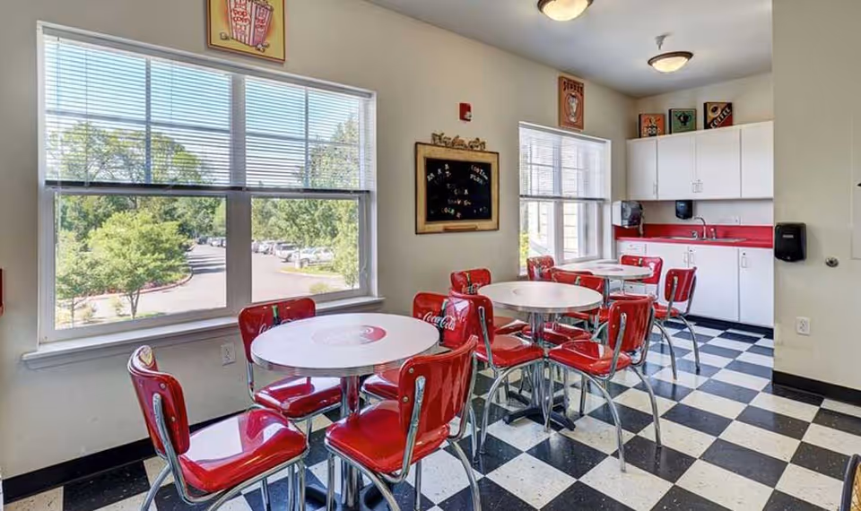 A bright dining area with large windows showing a view of trees and a parking lot outside. Inside, there are round white tables surrounded by red Coca-Cola branded chairs. The floor has a black and white checkered pattern. The back wall features white cabinets with a red countertop, a sink, and a paper towel dispenser. There are vintage-style Coca-Cola posters on the walls above the windows and cabinets.