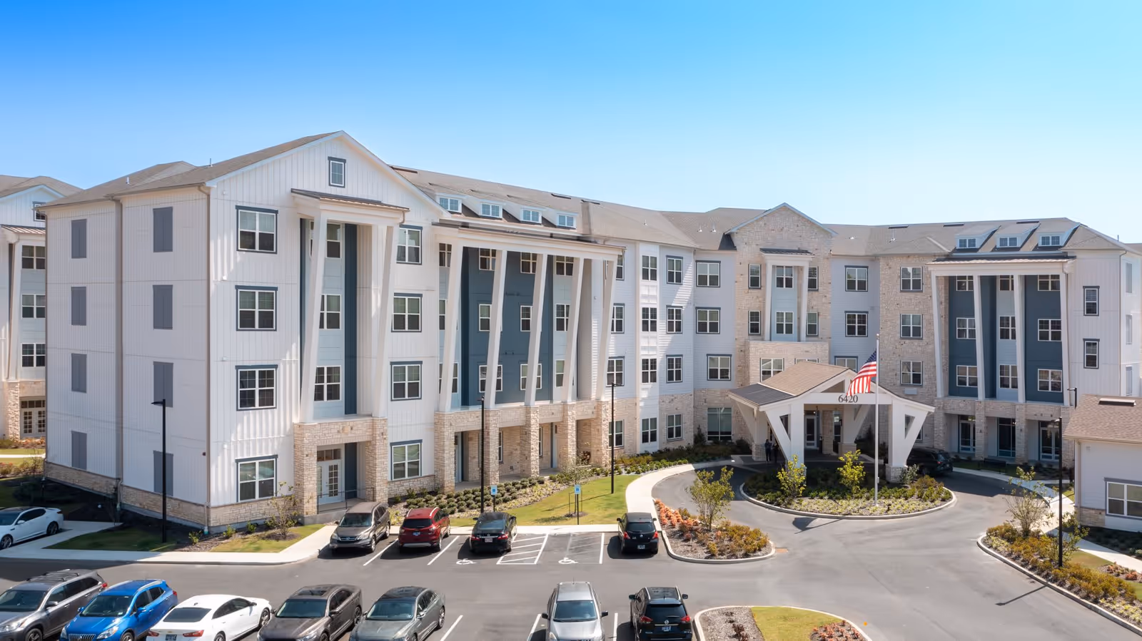 Exterior view of a large, modern senior living facility building with multiple floors, a covered entrance with an American flag, and a parking lot with several cars parked in front under a clear blue sky.