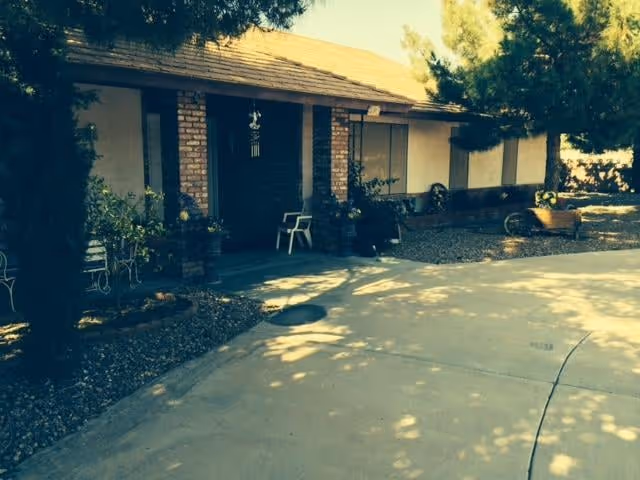 Exterior view of a single-story residential building with a covered porch supported by brick columns. The porch has a white chair and some potted plants. The driveway and surrounding landscaping with trees and shrubs are visible in the foreground.