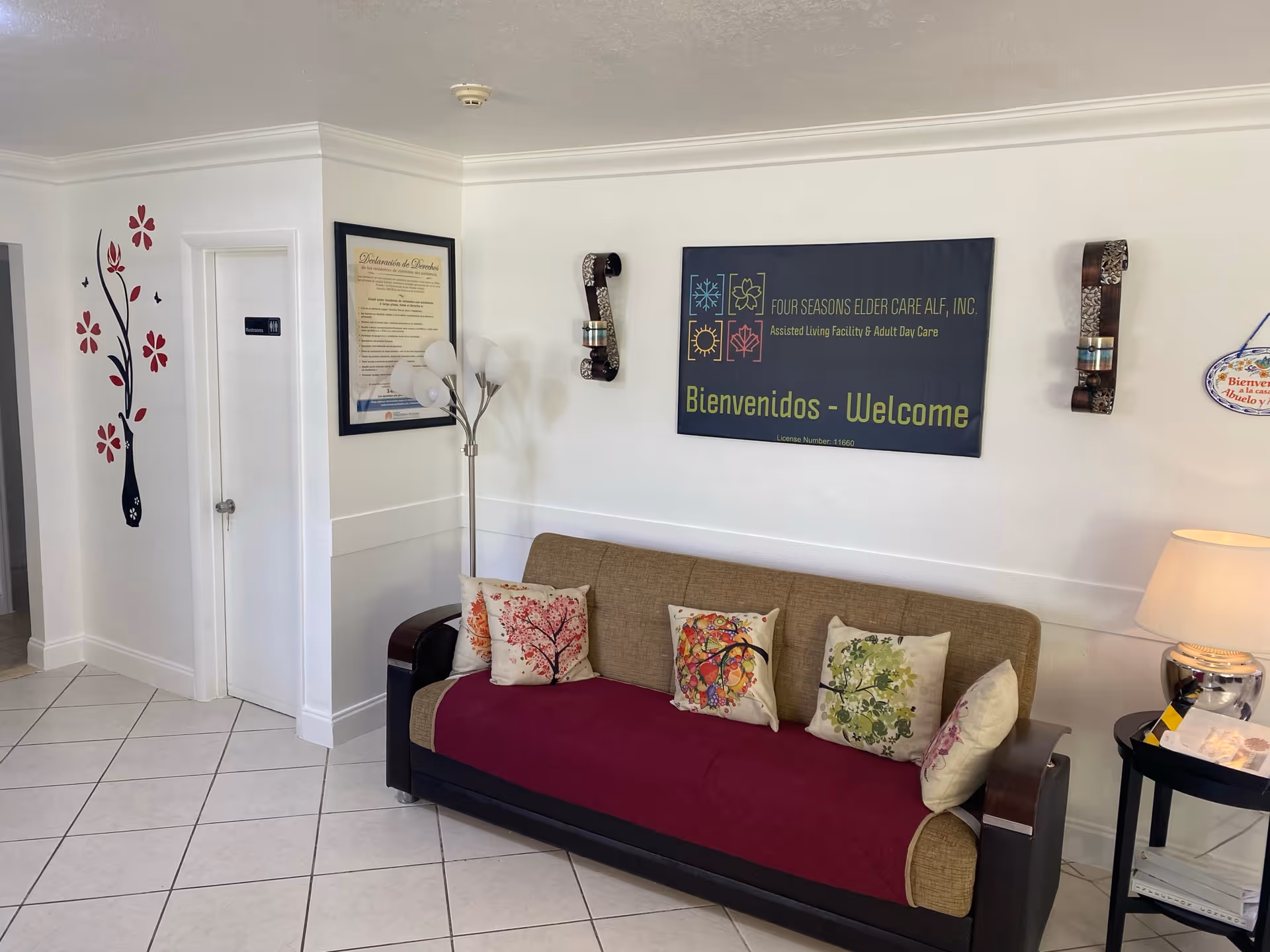 A cozy waiting area inside FOUR SEASONS ELDER CARE ALF, INC. featuring a beige sofa with colorful floral cushions and a red throw. Above the sofa is a welcoming sign with the facility's name and the words 'Bienvenidos - Welcome'. The walls are white with decorative wall sconces and a framed document. To the left, there is a door with a restroom sign and a wall decal of a tree with red flowers. A floor lamp stands next to the sofa, and a small round table with a lamp and some papers is on the right.