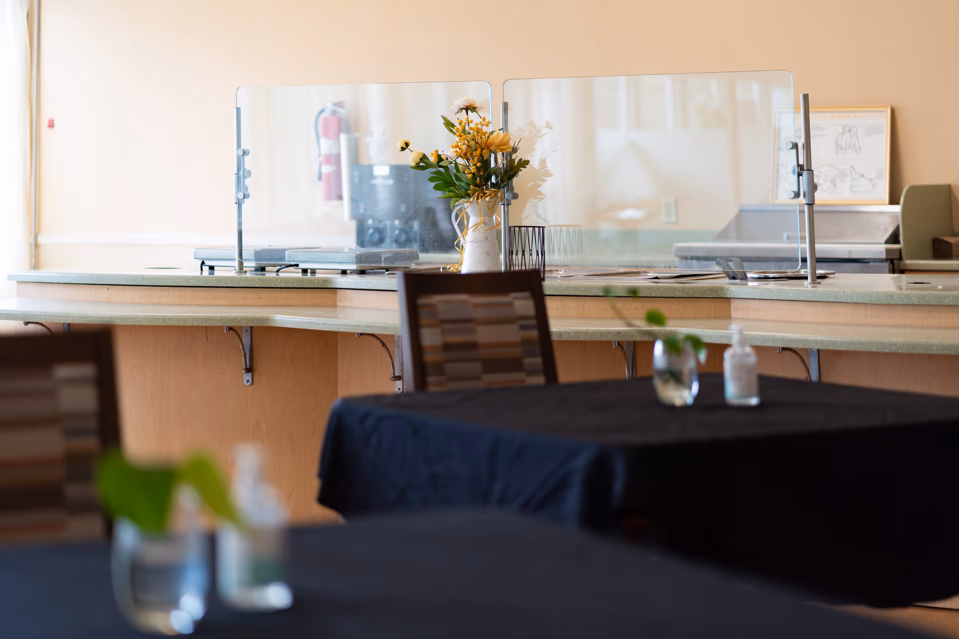 Interior view of a dining area with tables covered in black tablecloths, each table having a small plant and a bottle of hand sanitizer. In the background, there is a counter with a clear protective barrier and a vase with yellow flowers. The setting appears clean and organized.