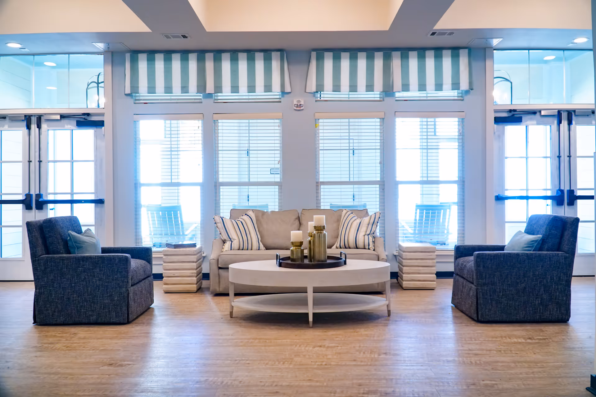 A bright and airy living room with a beige sofa adorned with striped pillows, two dark blue armchairs with light blue cushions, a round white coffee table with decorative candles, and large windows with striped valances letting in natural light.