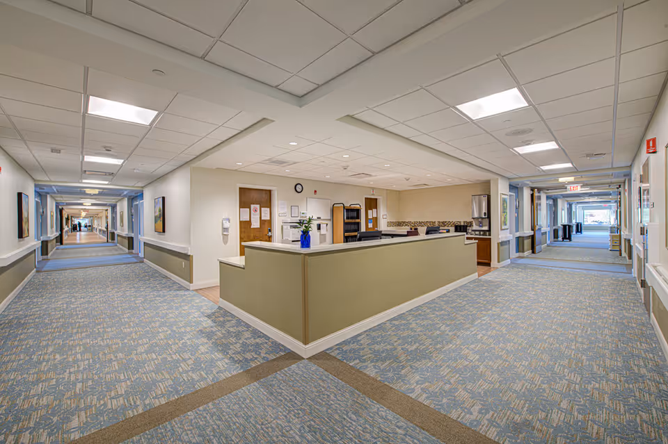 A wide hallway in a senior living facility with a central nurse's station featuring a green counter, office chairs, and a blue vase with flowers. The hallway has patterned blue and beige carpeting, white walls with handrails, ceiling lights, and doors along both sides. Artwork is hung on the walls, and there is an exit sign visible at the far end.