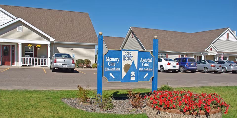 Exterior view of Our House Senior Living - Rice Lake Assisted Care facility showing a parking lot with several cars, a blue sign indicating directions to Memory Care and Assisted Care, and a flower bed with red flowers in the foreground.