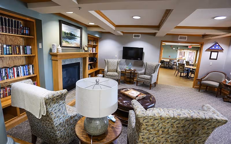 Cozy common room with upholstered chairs and a lamp arranged around a central ottoman, a fireplace with built-in bookshelves, and a wall-mounted TV opening into a dining area.