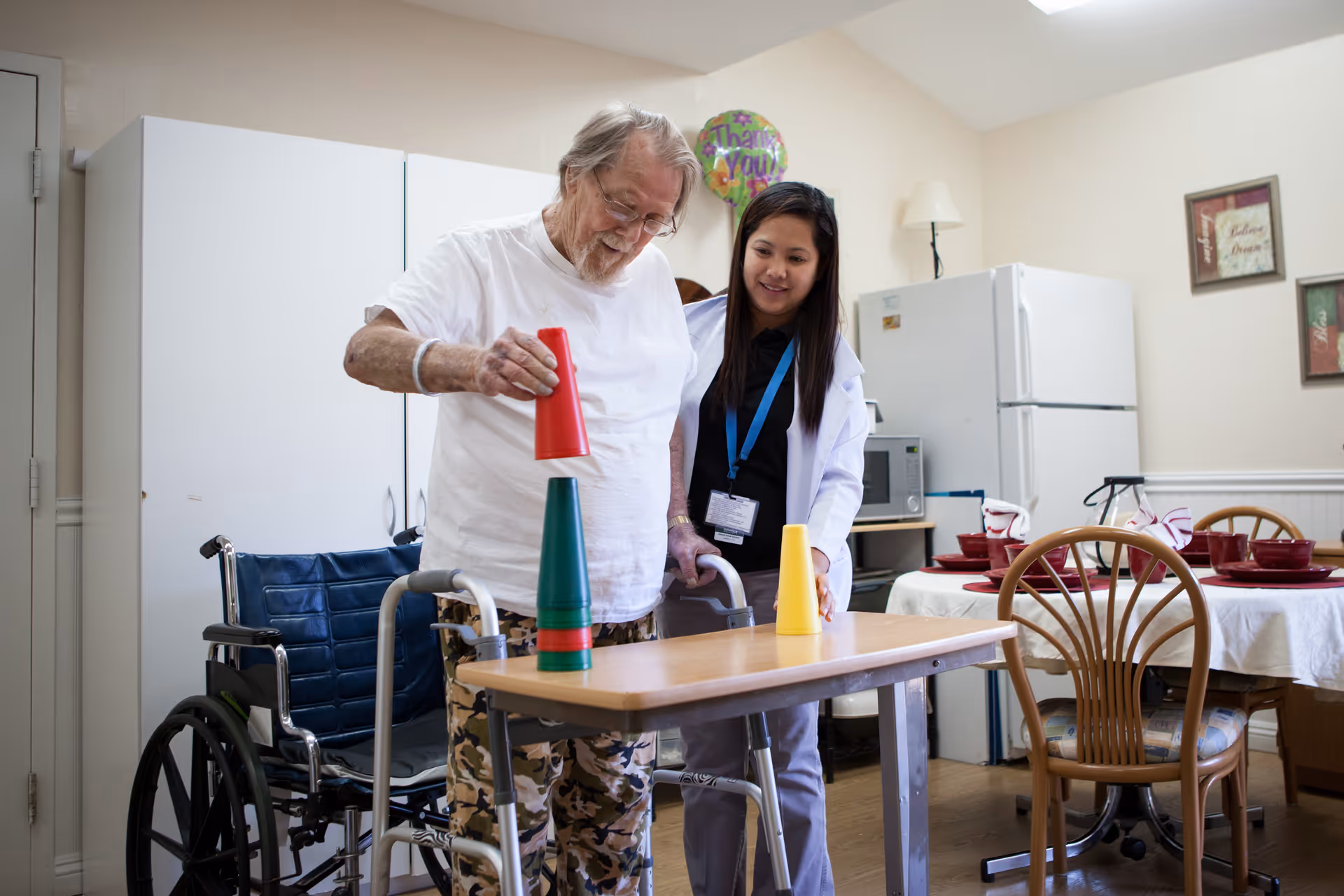 An elderly man using a walker is stacking colorful plastic cups on a table while a female caregiver stands beside him, smiling. In the background, there is a wheelchair, a refrigerator, a microwave, and a dining table set with red dishes and napkins. The room appears to be a common area in a senior living facility.