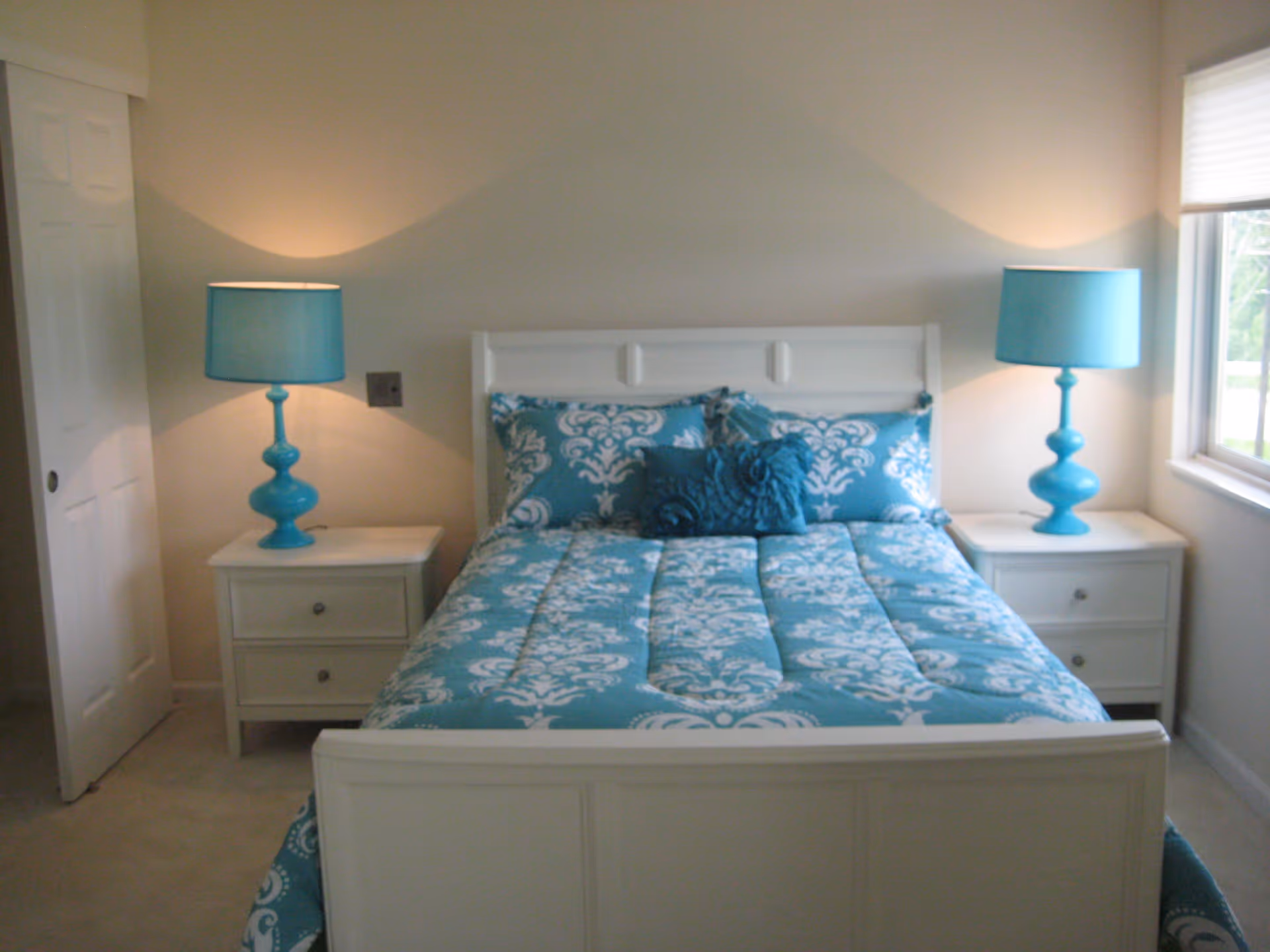 A bedroom with a white wooden bed frame and a blue and white patterned comforter and pillows. There are two matching white nightstands on either side of the bed, each with a blue lamp. A window with a white blind is on the right side of the room, and a white sliding door is on the left.