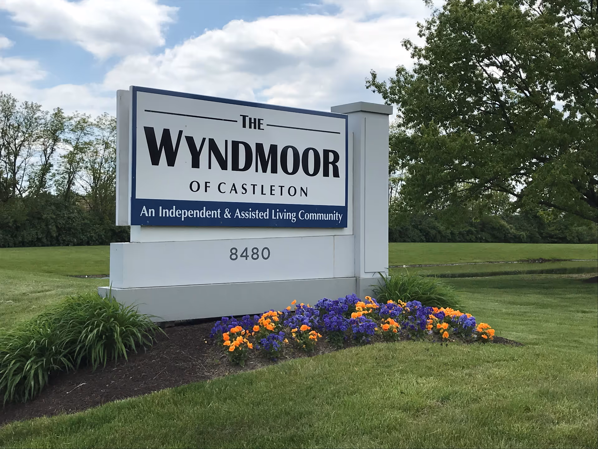 Outdoor sign for The Wyndmoor of Castleton, an independent and assisted living community, surrounded by green grass, colorful flowers, and trees under a partly cloudy sky.