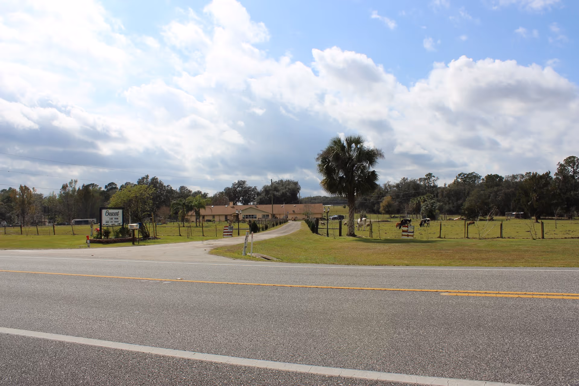 Wide exterior view of Crescent Lake House showing a driveway leading to a low building, fenced pastures with grazing horses and a roadside sign.