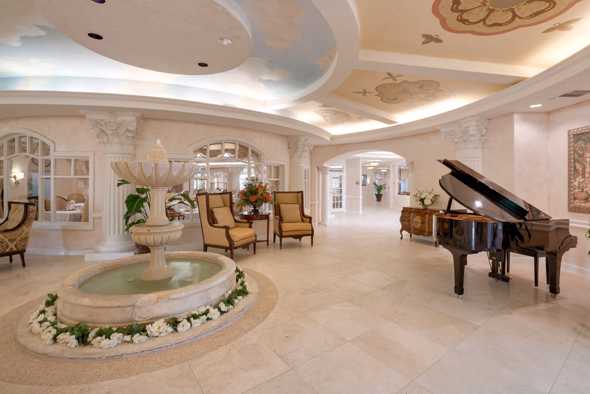 Elegant senior living facility interior with a central stone water fountain surrounded by white flowers, two upholstered armchairs with a small table and flower arrangement, a black grand piano, decorative columns, and a ceiling painted with clouds and floral designs.