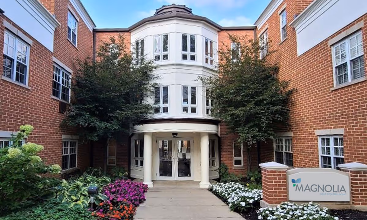 Exterior view of a brick senior living facility building with white framed windows, a central entrance with white columns, and landscaped flower beds with colorful flowers and green shrubs on either side of the walkway. A sign in front reads 'MAGNOLIA DIAL SENIOR LIVING'.