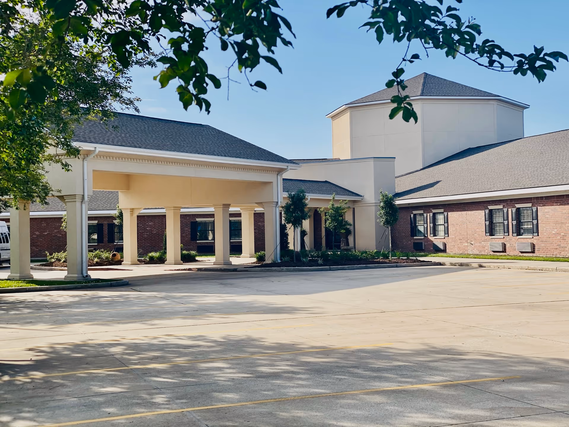 Exterior view of a senior living facility building with a covered entrance supported by columns, surrounded by small trees and landscaping. The building has a combination of brick and light-colored walls with multiple windows and a large octagonal structure in the background. The foreground shows an empty parking lot with yellow lines marking parking spaces.