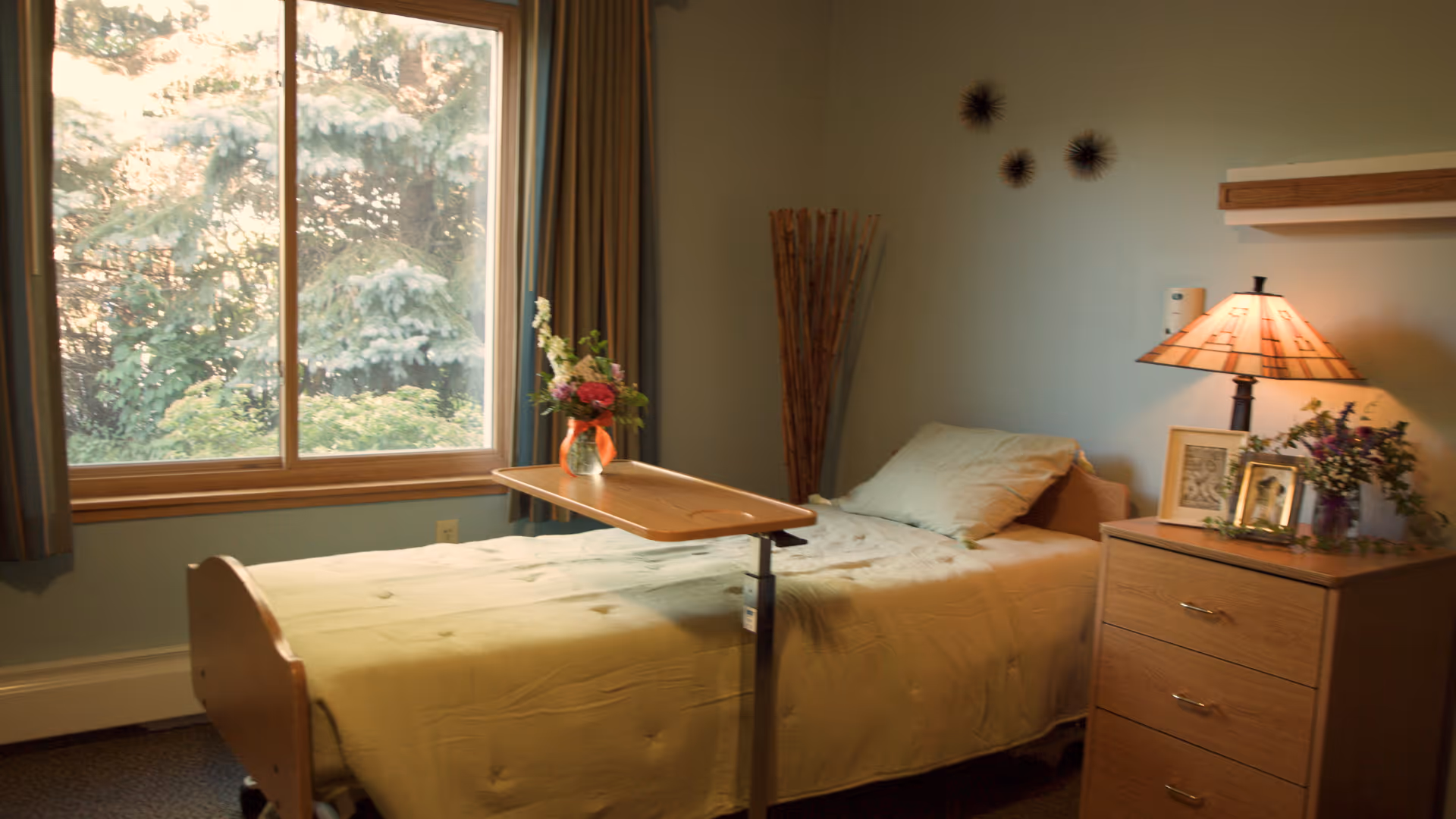 A simple furnished bedroom with a single bed, overbed table holding a vase of flowers, a bedside dresser and lamp, and a large window showing trees outside.