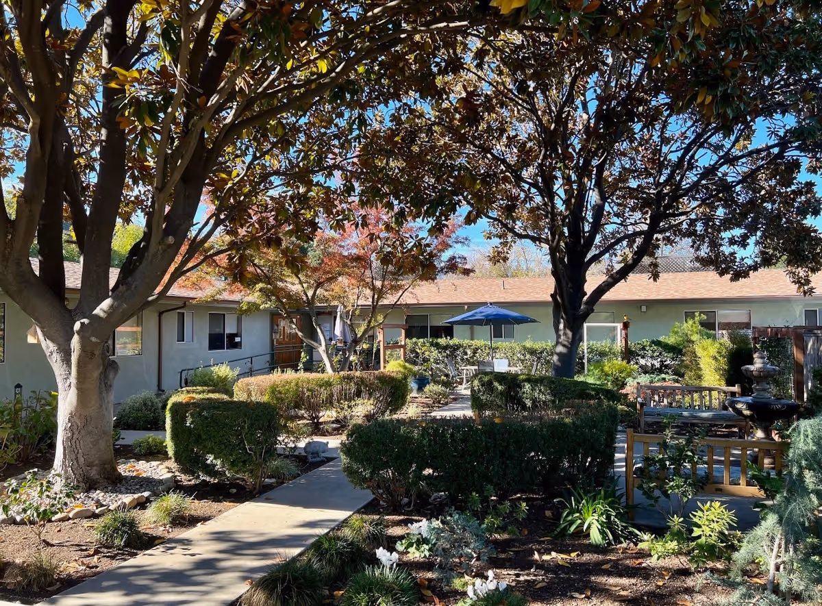 A peaceful outdoor garden area at Cedar Creek Alzheimer's and Dementia Care facility featuring a paved walkway, trimmed bushes, trees with autumn leaves, a blue patio umbrella, benches, and a water fountain under a clear blue sky.