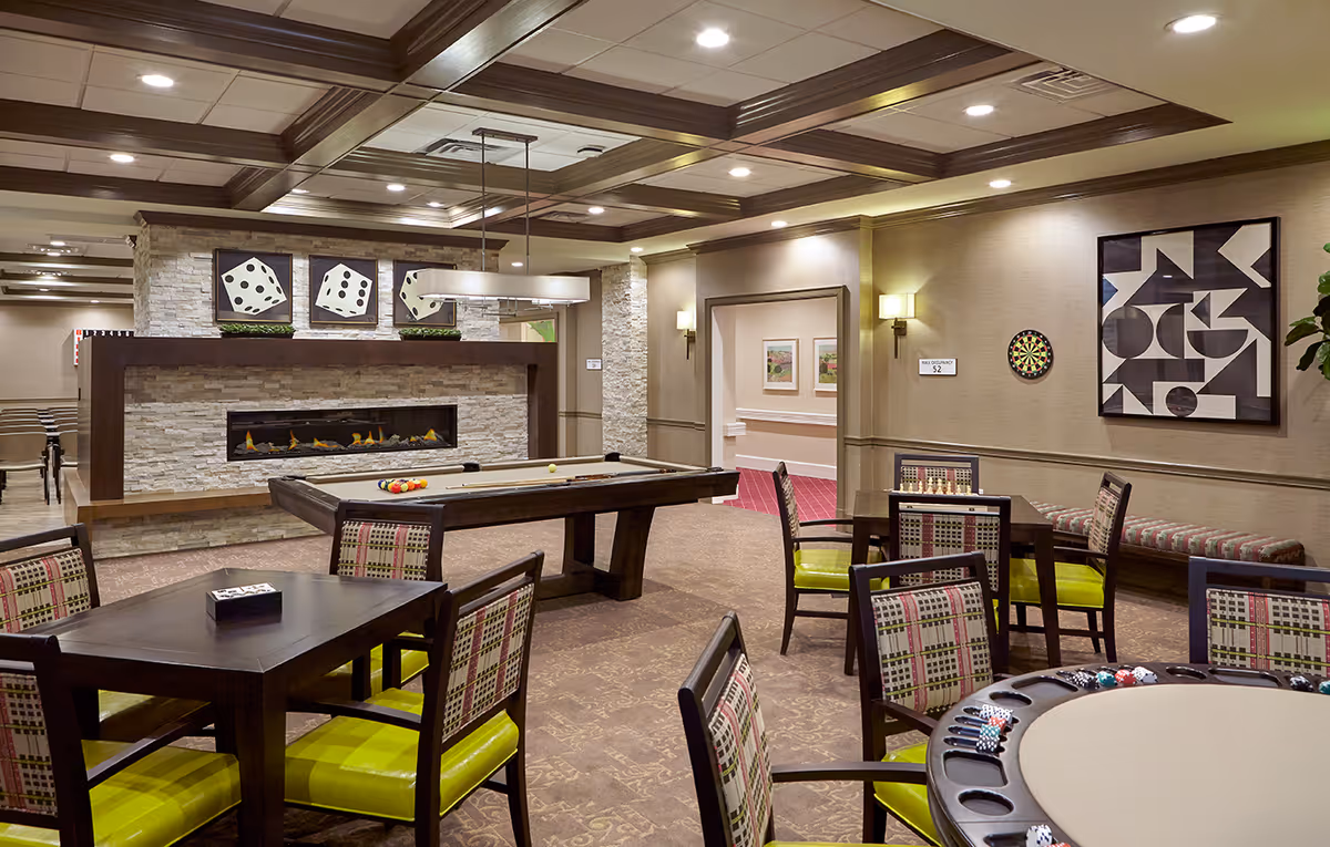 A spacious recreational room in an assisted living facility featuring a pool table, a poker table with chips, and several tables with chairs. The room has a modern fireplace with decorative dice artwork above it, a dartboard on the wall, and abstract art. The ceiling has wooden beams and recessed lighting, and the walls are decorated with sconces and framed pictures.