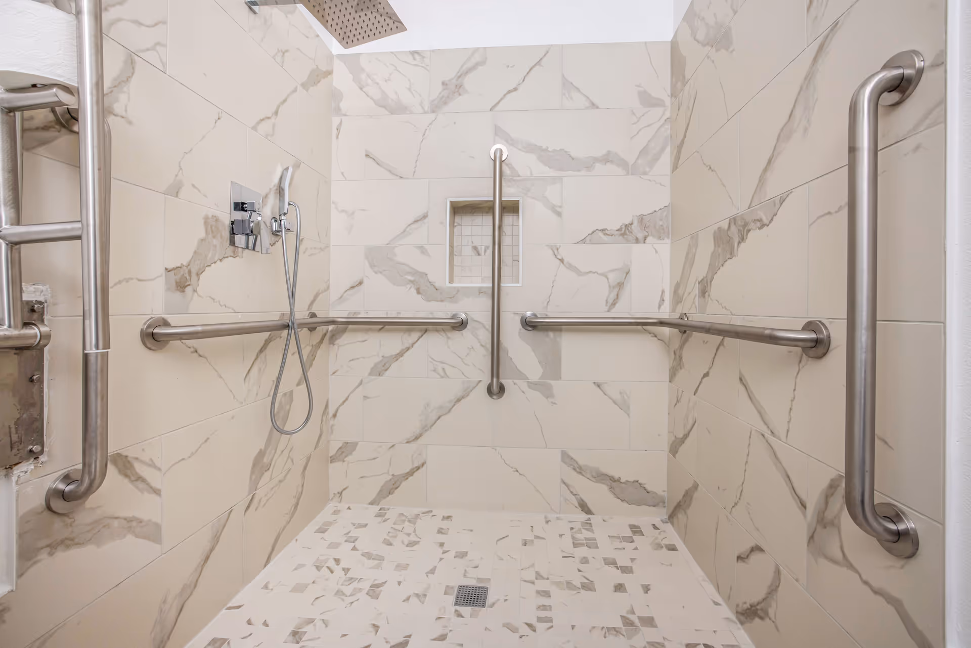 Accessible tiled walk-in shower with multiple stainless grab bars, a handheld showerhead, and a recessed niche.