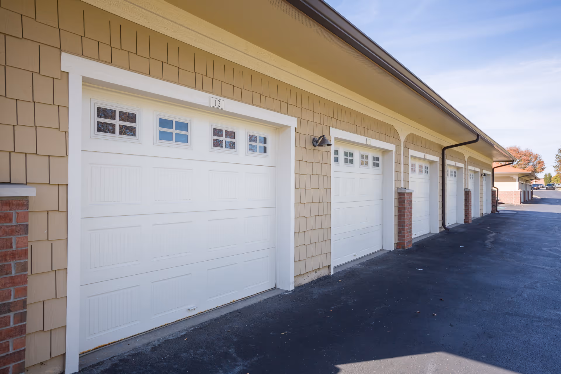 A row of white garage doors with small windows at the top, attached to a beige building with brick accents, under a clear blue sky.