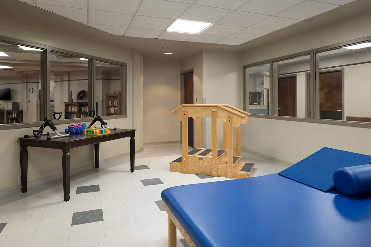 Interior view of a therapy room at St. Ann's Skilled Nursing & Therapy featuring a blue therapy table with a bolster pillow, a wooden set of stairs with handrails for physical therapy, and a table with various therapy equipment including hand weights and exercise tools. The room has beige walls, large windows looking into adjacent rooms, and a tiled floor with gray and white squares.