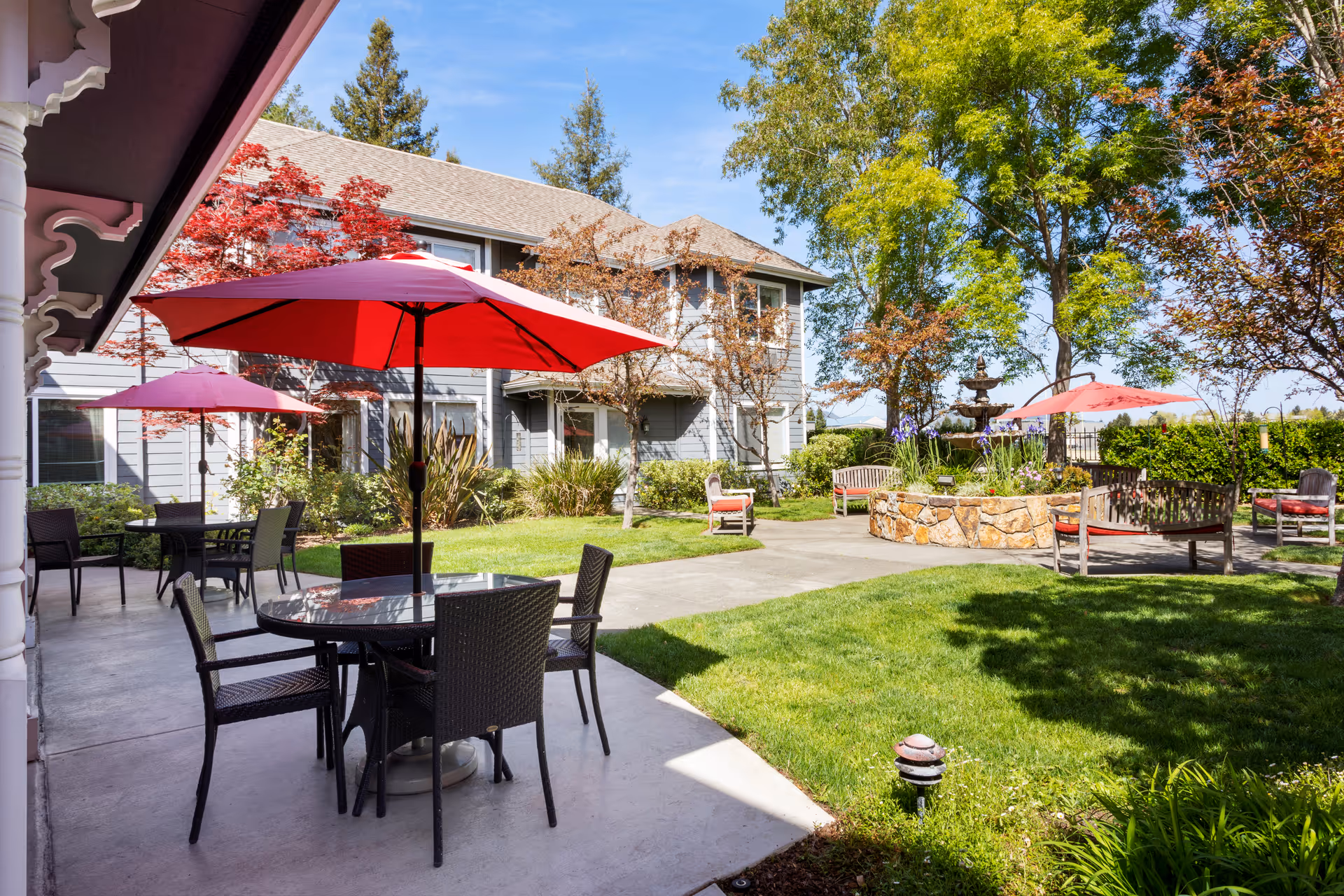 Outdoor patio area at Aegis Living Napa with round tables and chairs under red umbrellas, surrounded by green grass, trees, and shrubs. There are benches and a stone planter with a fountain in the background, and a two-story building with gray siding and windows behind the garden.