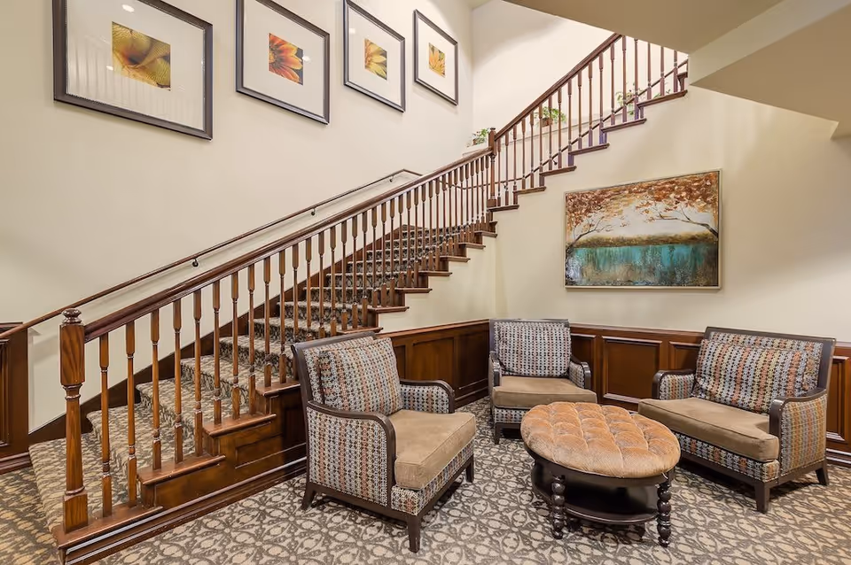 A cozy seating area in a senior living facility with two armchairs and a loveseat upholstered in patterned fabric, arranged around a round tufted ottoman. Behind the seating area is a wooden staircase with carpeted steps and a wooden railing. The walls are decorated with framed artwork and a large painting of a tree and water scene.