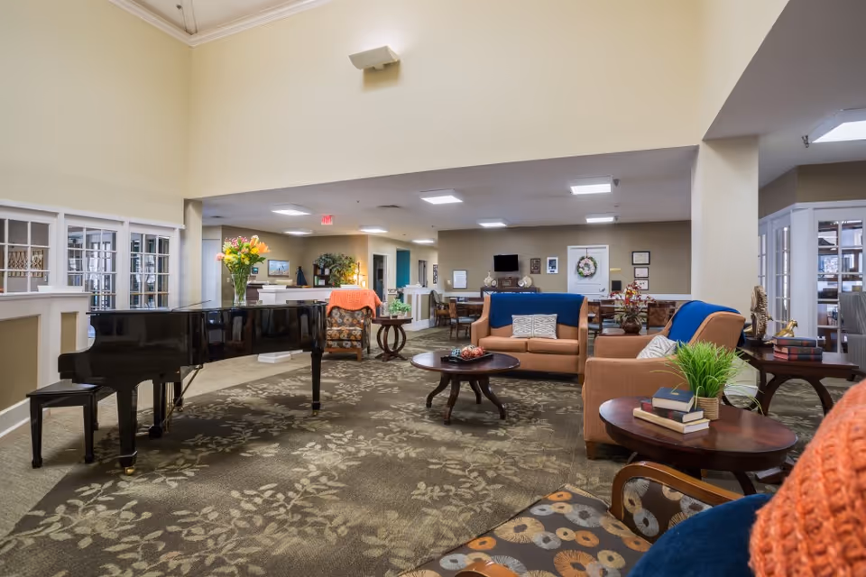 A spacious and well-lit living room area in a senior living facility featuring a black grand piano with a vase of flowers on top, several comfortable armchairs and sofas with cushions and throws, round wooden coffee tables with decorative items and books, and a patterned carpet. The walls are painted in neutral tones, and there are framed pictures and a television mounted on the far wall.